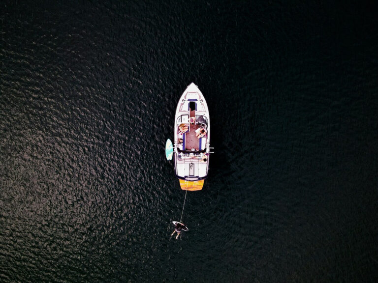 Aerial view of a boat with seasonal boat moorage on dark water, several people relaxing on deck, and one person floating on an inflatable nearby. Fill your empty slips fast with Marina Moorage