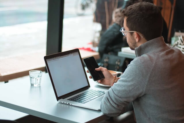 A person sits at a table in a cafe, using a smartphone and laptop—perhaps checking their marina management software. A glass of water and coffee cup are on the table. The person is facing away from the camera. Fill your empty slips fast with Marina Moorage