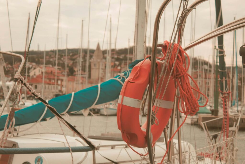 Orange lifebuoy with rope attached to a sailboat railing; marina with seasonal boat moorage, other boats, and a town with a church in the background. Marina Safety is a top priority, get tips from MarinaMoorage.com