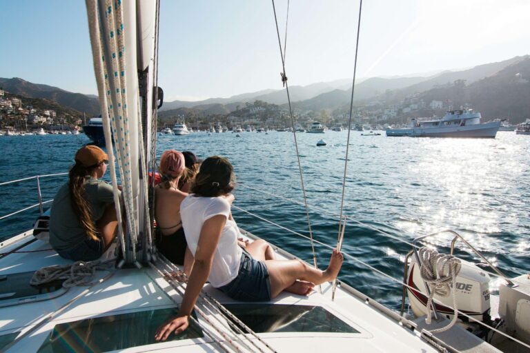 Three people relax on the deck of a sailboat, overlooking a harbor where boats fill marina slips, with hills in the background on a sunny day. Fill your empty slips fast with Marina Moorage