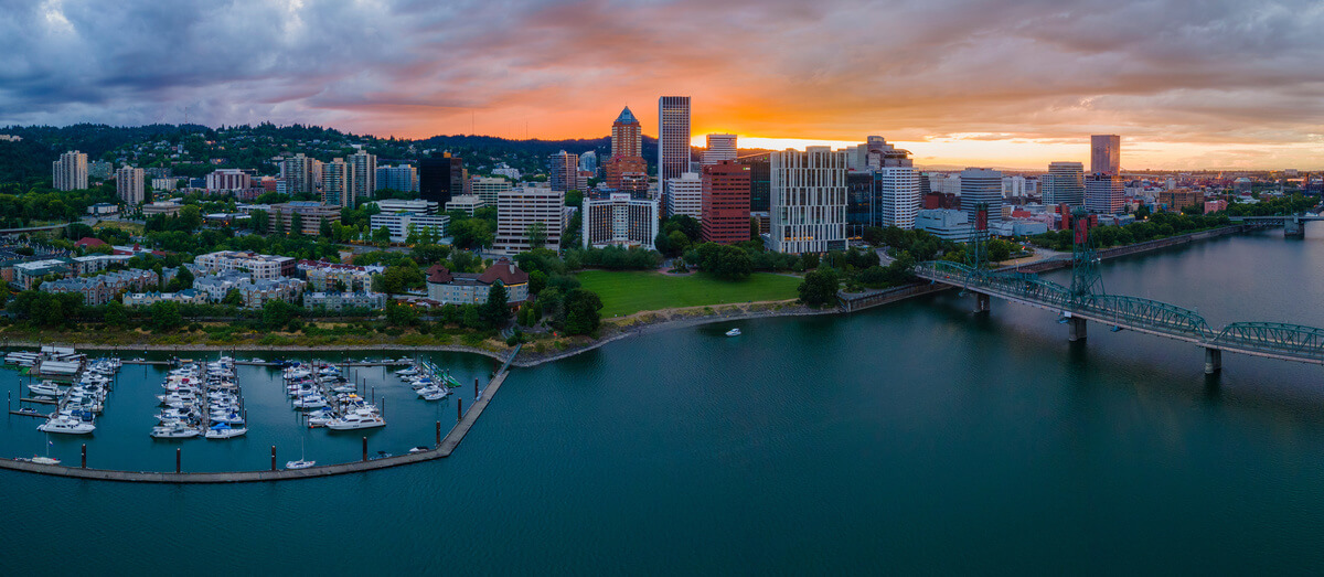 A panoramic view of a city skyline at sunset, with empty slips in the marina, a green park, tall buildings, and a bridge spanning a river in the foreground. Fill your empty slips fast with Marina Moorage