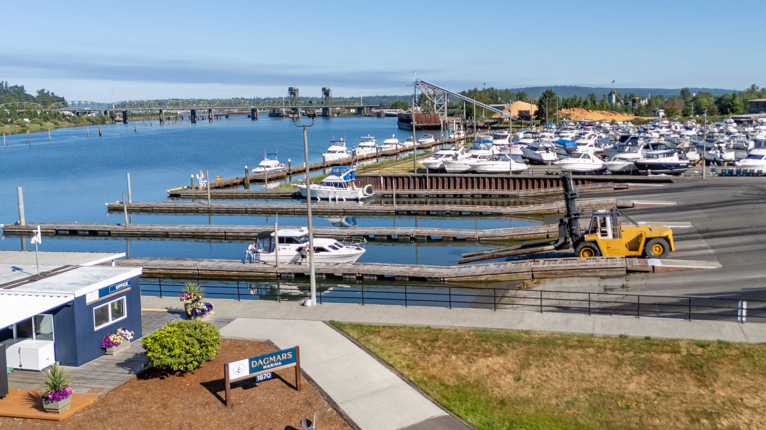 A yellow forklift moves a boat near the docks at Dagmars Marina, with several boats in the water and a wood chip pile in the background. Fill your empty slips fast with Marina Moorage
